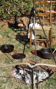 Photo of a metal tripod over a fire, with a large metal cookpot on the ground next to it
