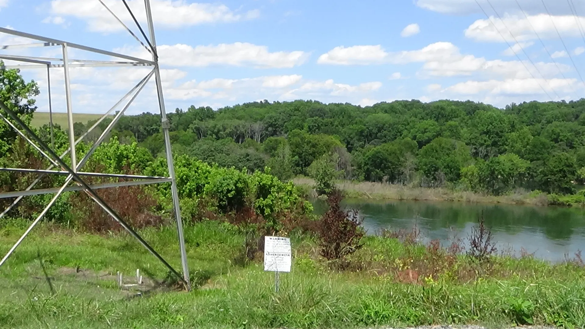 Photo of woods across a river, with the base of a transmission tower in the left foreground