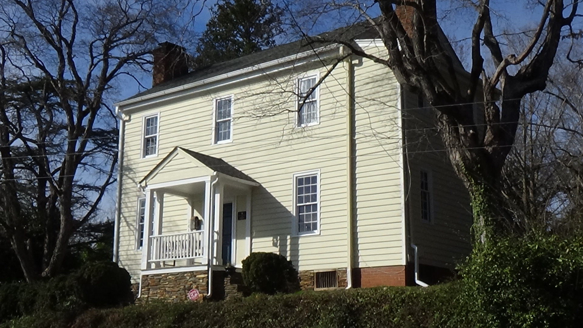 Photo of a two-story, wood-framed house atop an embankment, with a porch-covered entrance, painted in light yellow