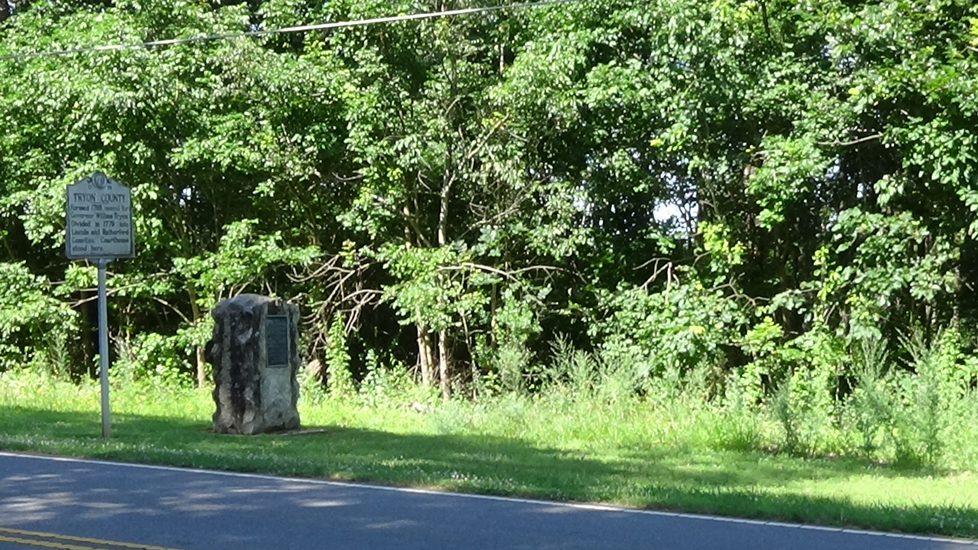 Photo of a granite monument and historical marker by a two-lane road with trees behind them