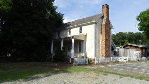 Photo of a two-story wooden house with a porch across part of the first floor, the left side obscured by a tree