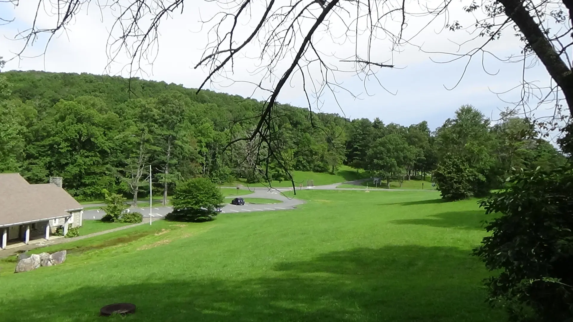 Photo of a large meadow sloping downhill to the left, with the small museum building on that edge and a highway in the background below a wooded rise
