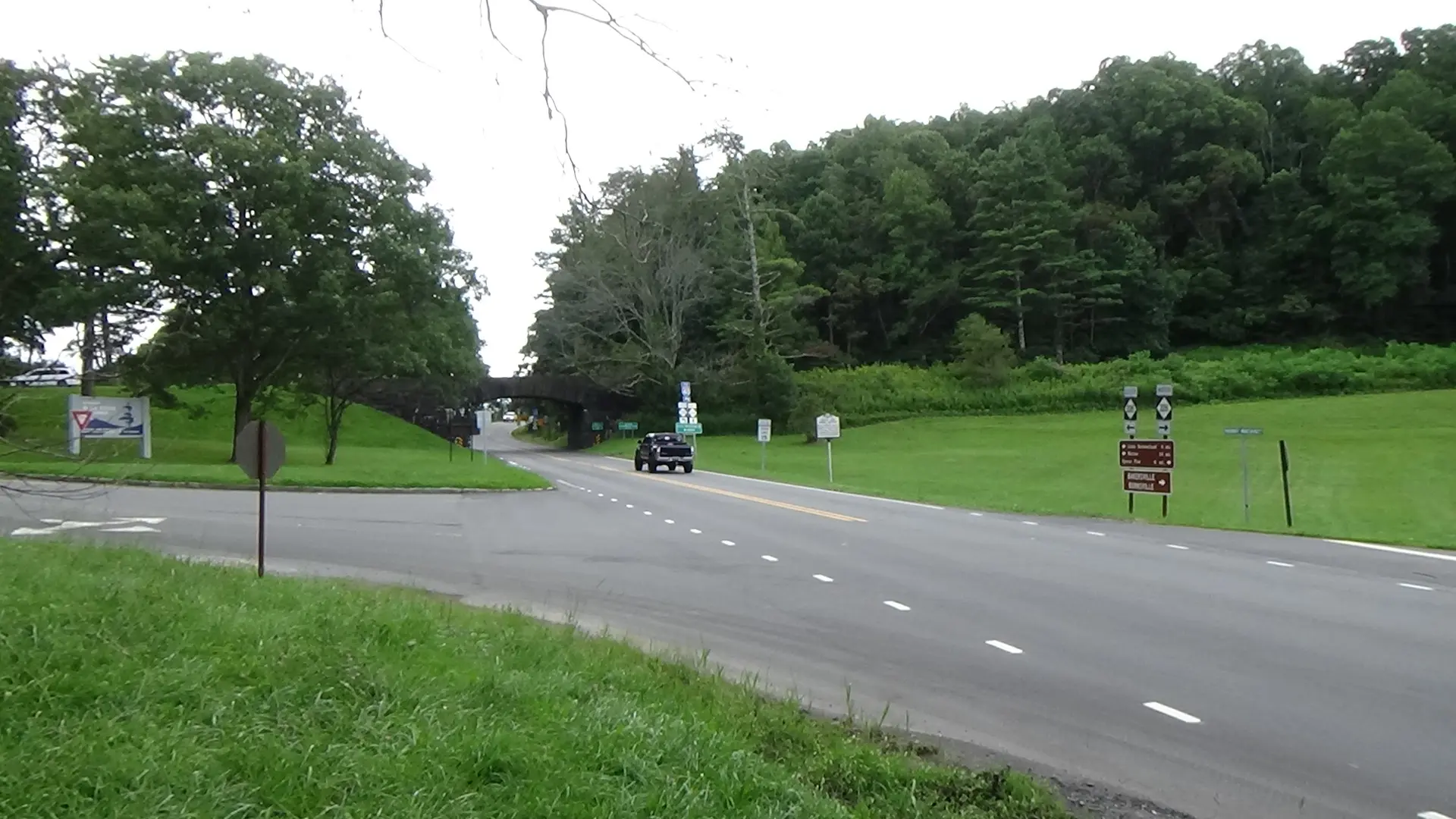Photo of a two-lane highway through grassy fields leading to the top of a tree-lined ridge, with an overpass visible there