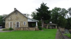 Photo of the Gateway Museum from the side, a stone one-story building with a pointed roof and porch, and a gazebo on the right with the river further right