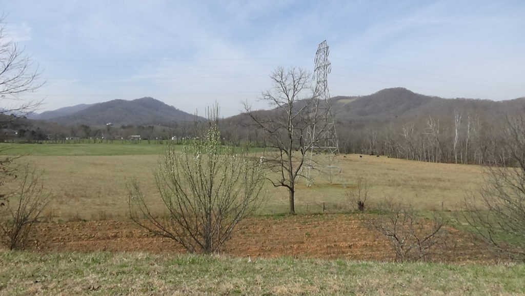 Photo of a green pasture with a plowed field in the front and mountains behind