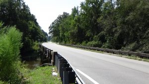 Photo of a flat bridge between guardrails carrying a two-lane road over a creek visible to the left, with trees lining both sides into the distance