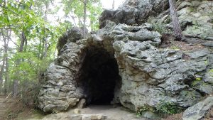 Photo of a narrow cave opening in a large outcrop of rock, with trees to the left