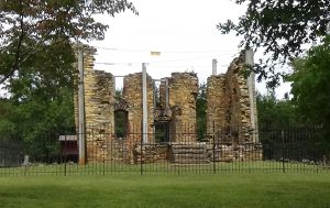 Photo of the roofless ruins of a house of light-brown rocks, with vertical stabilizing poles around it and a black security fence