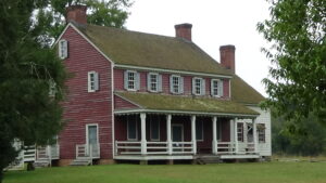 Photo of a two-story, red-painted wooden house with a first-floor porch and single-story wing off the right end