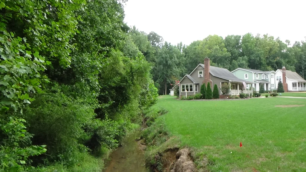 Photo of lawn and houses on right, creek and trees on the left, and trees behind