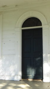 Photo of black door from the outside with a half-moon window above, and a half-dozen bullet holes in the white wall to the left