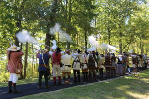 Photo of re-enactors in frontier clothing, seen from the back, on a battlefield trail firing their muskets