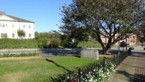 Photo of a triangular lawn within a low black metal fence, with a low monument on the right