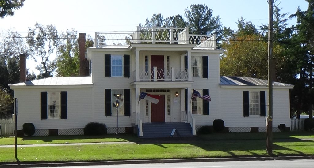 Photo of a white, wood, home, two stories in the main section with two one-story wings, and porches in the center upstairs and down