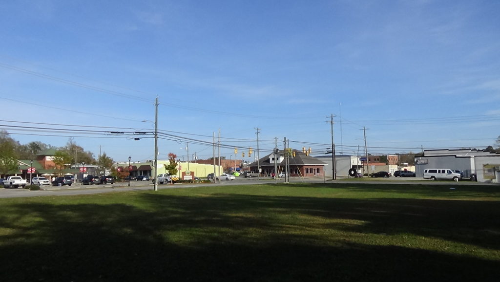 Photo of an empty, grass-covered lot looking toward a small-town intersection with low buildings along the horizon