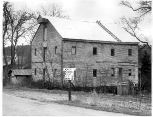 Black and white photo of an old, two-and-a-half-story, wooden building by a road, four-windows deep, with doors at each level in the front