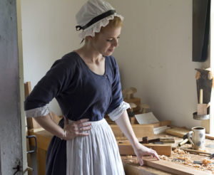 A woman in a 1700s blue dress, white apron, and white bonnet standing at a carpenter's bench