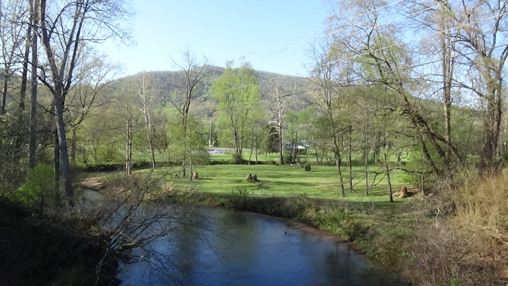 A park seen behind a river flowing away from the camera, with a mountain further behind