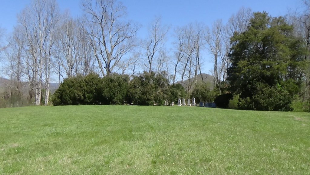 A small cemetery surrounded by large bushes on a grass-covered rise, with leafless trees and a blue sky behind.