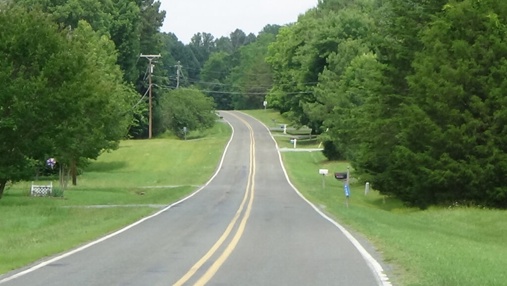 Photograph of a two-lane road running downhill between yards and trees