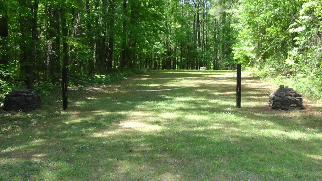 Photo of rectangle of grass stretching away from the camera, surrounded by trees, with round monuments on either side of a chain gate and another in the distance