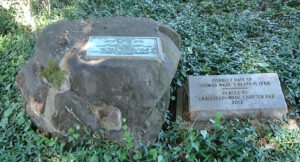 Photo of a rock about two feet high with a plaque on it, and another plaque to its right about the grave